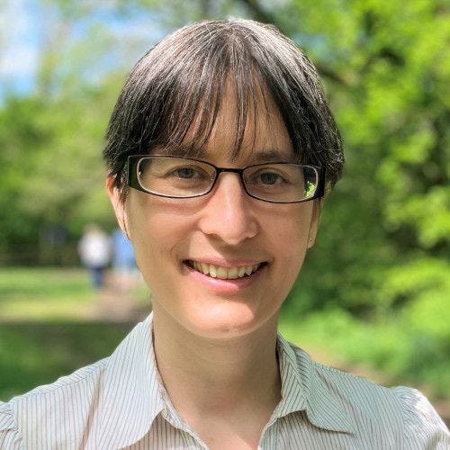 Heather Turner outside in the sun, smiling. She is a white woman in her 40s with dark straight hair tied back and wears glasses. She is dressed in a business casual shirt.