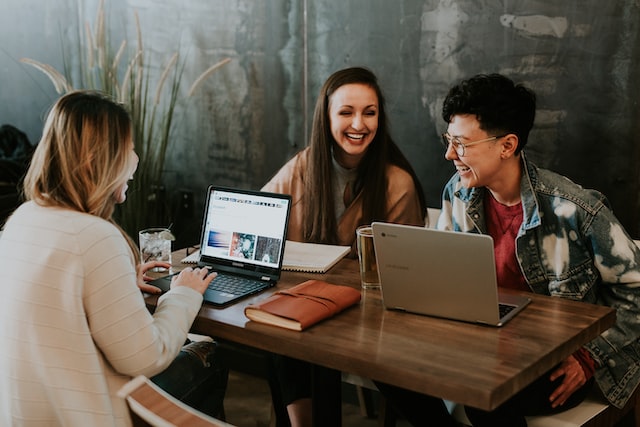 Three people laughing together as they sit around a table with laptops, notebooks and drinks.