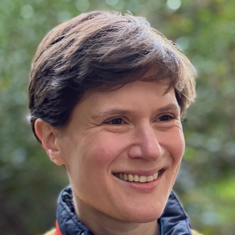 Headshot of Ella outside in the sun, smiling, looking slightly off-camera. She is a white woman in her 40s with short dark hair.