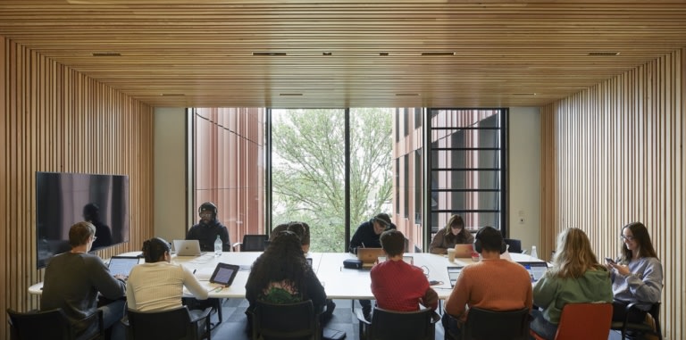 A group of 11 people around a long table with a large screen at one end and a big window behind. People are general busy working on laptops, writing on notepads, etc.